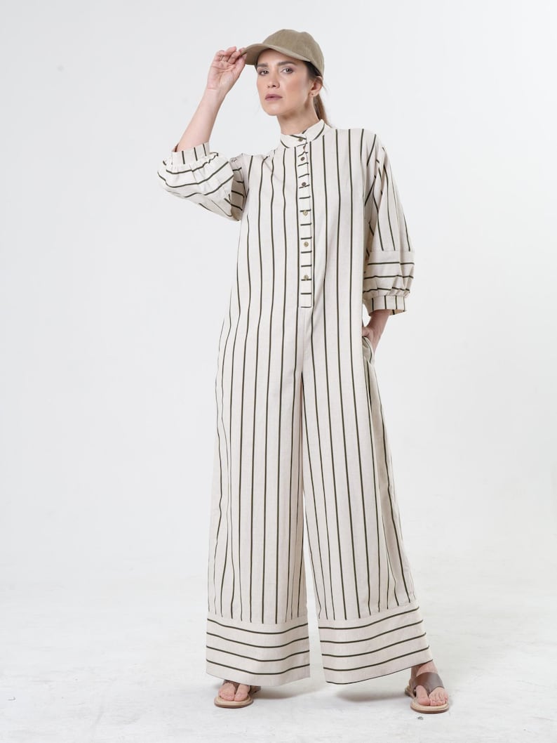 May include: A white and black striped jumpsuit with a button-up front and long sleeves. The jumpsuit has a wide leg and a relaxed fit. The model is wearing a light brown cap.