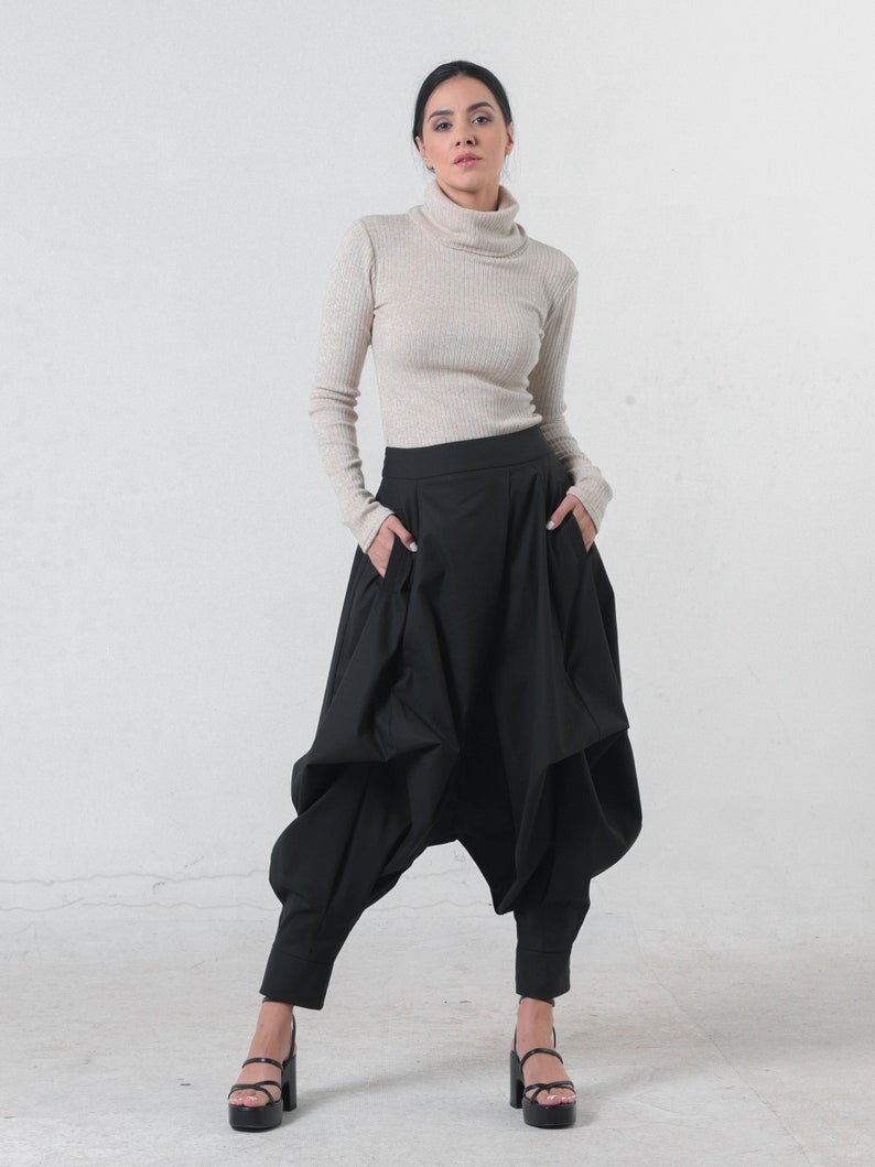 May include: A woman wearing a beige turtleneck sweater and black, loose-fitting pants with a unique, gathered design. She is wearing black platform sandals.