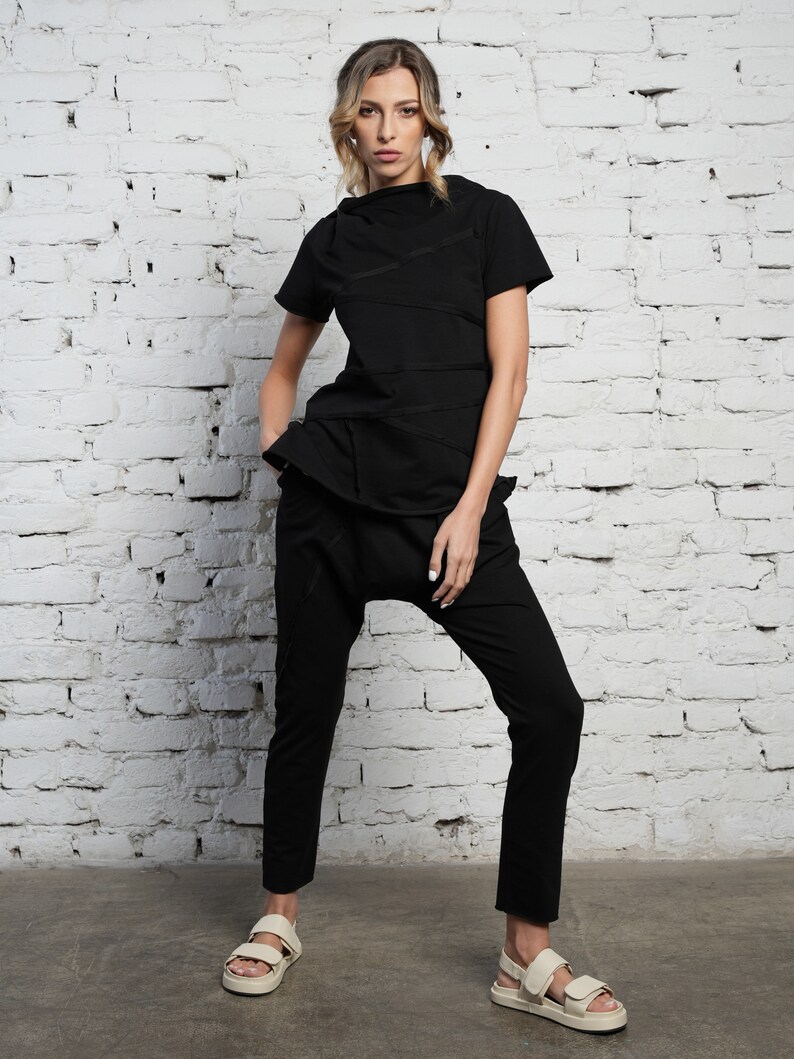May include: A black mock neck shirt with a layered design and black pants. The shirt has multiple zippers along the seams. The person is wearing white sandals.