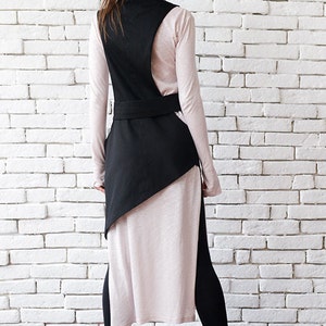 May include: A black asymmetrical vest with a high neck and a belt. The vest is worn over a long-sleeved, light pink shirt and black leggings.