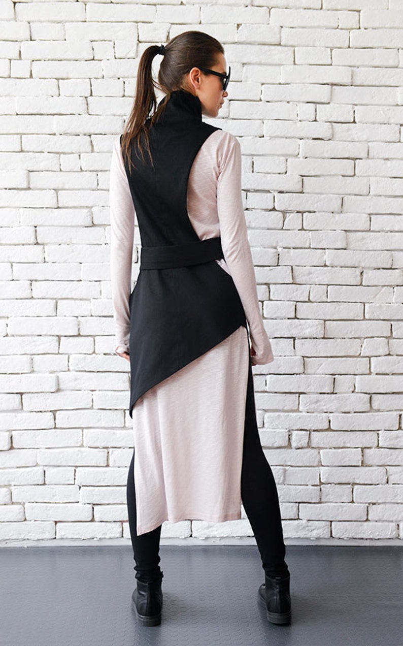 May include: A black asymmetrical vest with a high neck and a belt. The vest is worn over a long-sleeved, light pink shirt and black leggings.