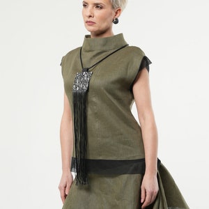 Puede incluir: Conjunto de top y falda de lino verde oliva. El top tiene cuello alto y detalles de malla negra. Un collar negro con colgante cuadrado y flecos. La falda tiene un dise&ntilde;o en capas. Pendientes negros completan el look.