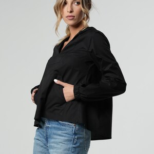 Puede incluir: Una camisa negra de manga larga con cuello en V y capucha con cord&oacute;n. La camisa tiene un corte holgado y un dobladillo ligeramente recortado. La modelo lleva la camisa con jeans azules.
