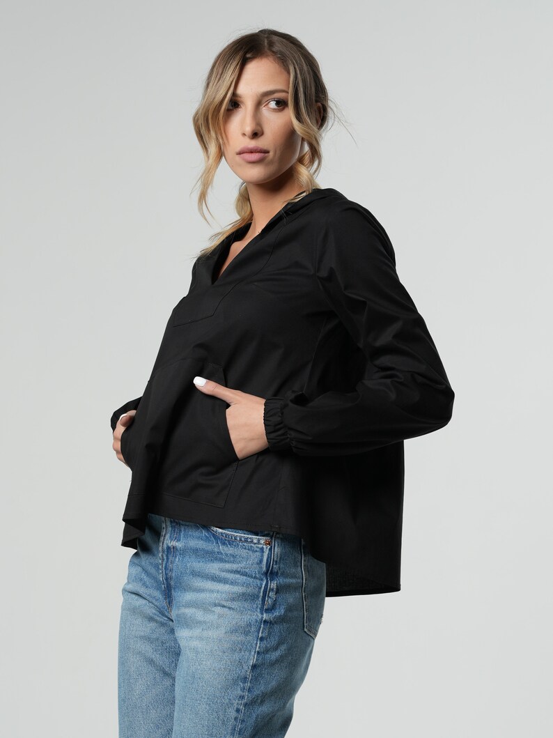 Puede incluir: Una camisa negra de manga larga con cuello en V y capucha con cord&oacute;n. La camisa tiene un corte holgado y un dobladillo ligeramente recortado. La modelo lleva la camisa con jeans azules.