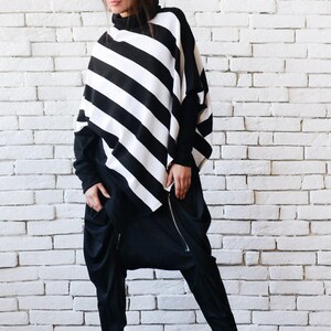 May include: A black and white striped poncho with a turtleneck collar and long sleeves. The poncho is worn over black pants with a zipper detail. The person is standing in front of a white brick wall.