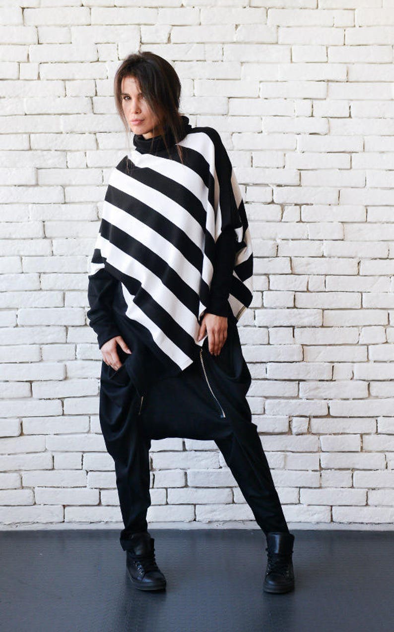 May include: A black and white striped poncho with a turtleneck collar and long sleeves. The poncho is worn over black pants with a zipper detail. The person is standing in front of a white brick wall.