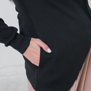 May include: Black ribbed knit top with a long sleeve and a diagonal hemline. The top has a pocket on the left side.