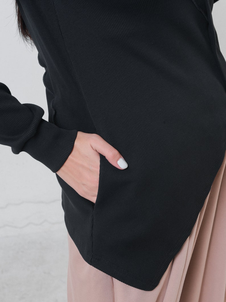May include: Black ribbed knit top with a long sleeve and a diagonal hemline. The top has a pocket on the left side.