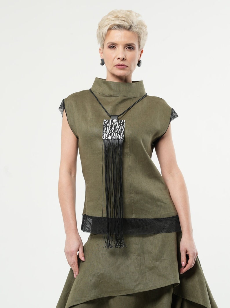 Puede incluir: Conjunto de top y falda de lino verde oliva. El top tiene cuello alto y detalles de malla negra. Un collar negro con un colgante rectangular y flecos es el punto focal. El colgante tiene texto. Pendientes negros.