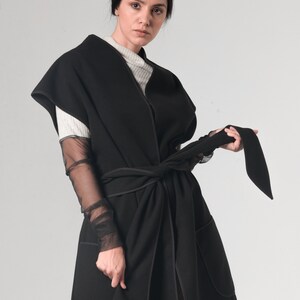 May include: A black sleeveless coat with a long, flowing silhouette. The coat has a wide, self-tie belt and a contrasting black stitching detail. The coat is made of a soft, comfortable fabric.