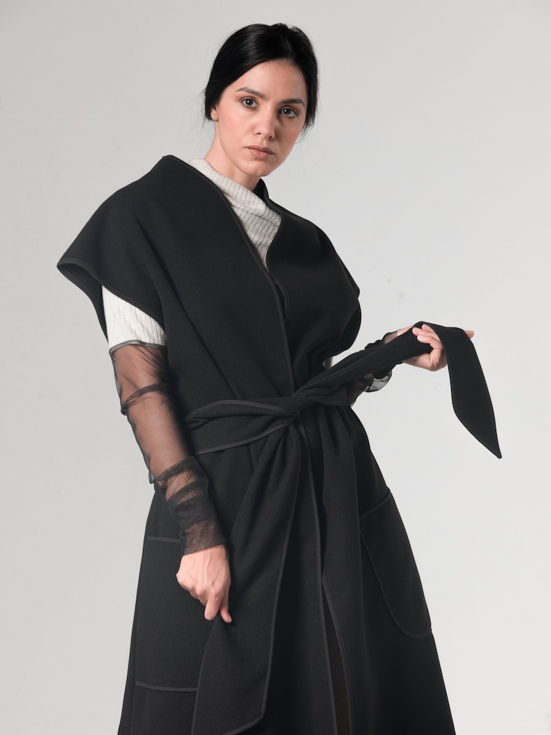 May include: A black sleeveless coat with a long, flowing silhouette. The coat has a wide, self-tie belt and a contrasting black stitching detail. The coat is made of a soft, comfortable fabric.