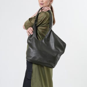 May include: A black leather tote bag with a curved top and a single strap. The bag is being held by a person wearing a green shirt.