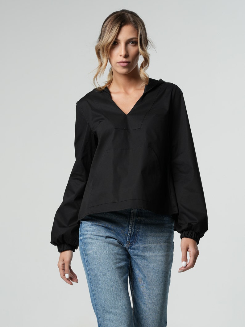 Puede incluir: Una blusa negra de manga larga con cuello en V y un detalle fruncido en las mu&ntilde;ecas. La blusa se lleva con pantalones vaqueros azules.