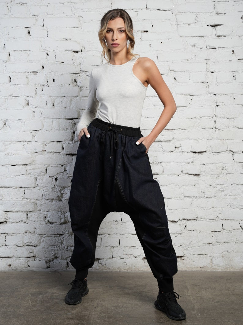 May include: A woman wearing a light gray one-shoulder top and dark blue denim pants with a unique, loose fit. She is standing in front of a white brick wall.