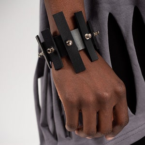 Puede incluir: Un brazalete de cuero negro con detalles met&aacute;licos plateados y tachuelas. El brazalete est&aacute; hecho de varias tiras de cuero que est&aacute;n conectadas entre s&iacute;.