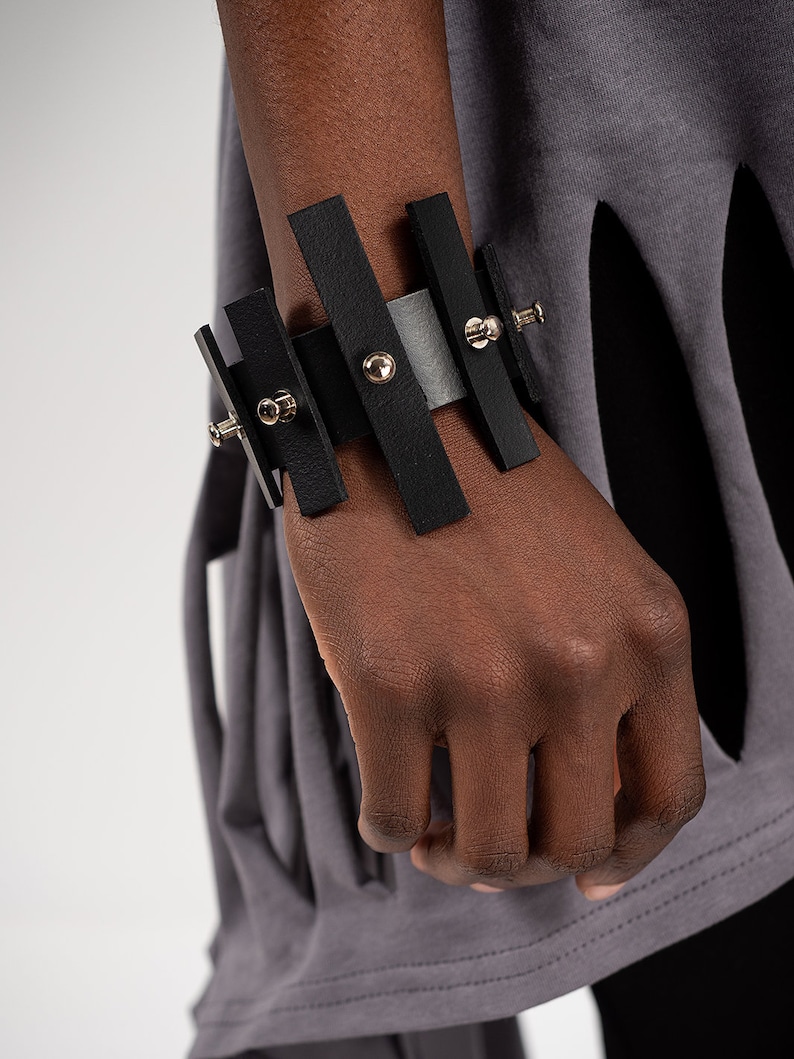 Puede incluir: Un brazalete de cuero negro con detalles met&aacute;licos plateados y tachuelas. El brazalete est&aacute; hecho de varias tiras de cuero que est&aacute;n conectadas entre s&iacute;.