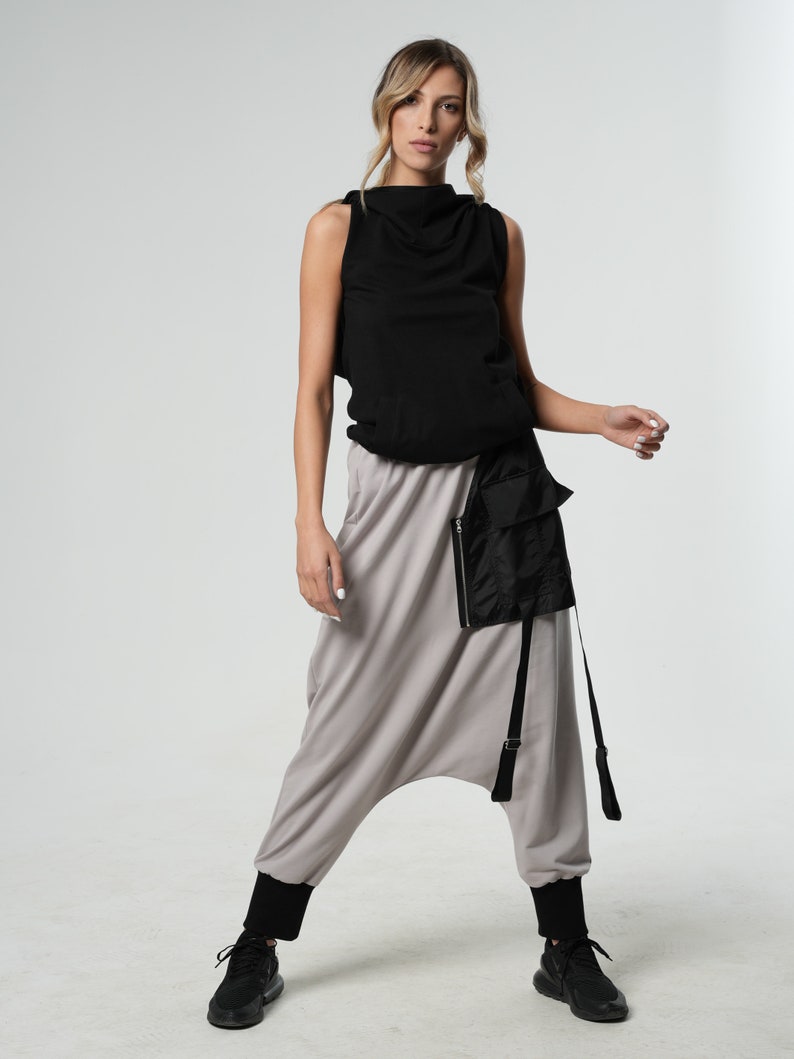 May include: A woman wearing a black sleeveless top with a high neckline and a pair of gray harem pants with a black utility belt and straps. She is wearing black sneakers.