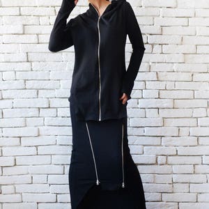 May include: A black hooded sweatshirt with a zipper and a matching black skirt with zipper details. The model is wearing black sneakers.