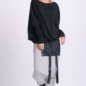 May include: A woman wearing a black long-sleeve top and gray harem pants with black accents. The pants have a black panel with a zipper and straps. Black shoes complete the outfit. The clothing is modern and stylish.