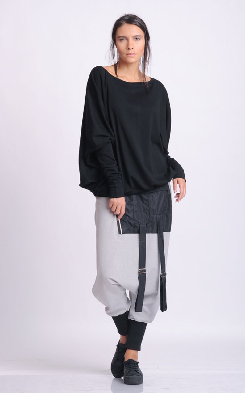 May include: A woman wearing a black long-sleeve top and gray harem pants with black accents. The pants have a black panel with a zipper and straps. Black shoes complete the outfit. The clothing is modern and stylish.