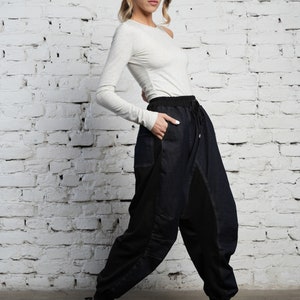 May include: A woman wearing a light gray long-sleeved shirt and dark blue denim pants with black panels. The pants have a drawstring waist and a relaxed fit. She is wearing black sneakers.
