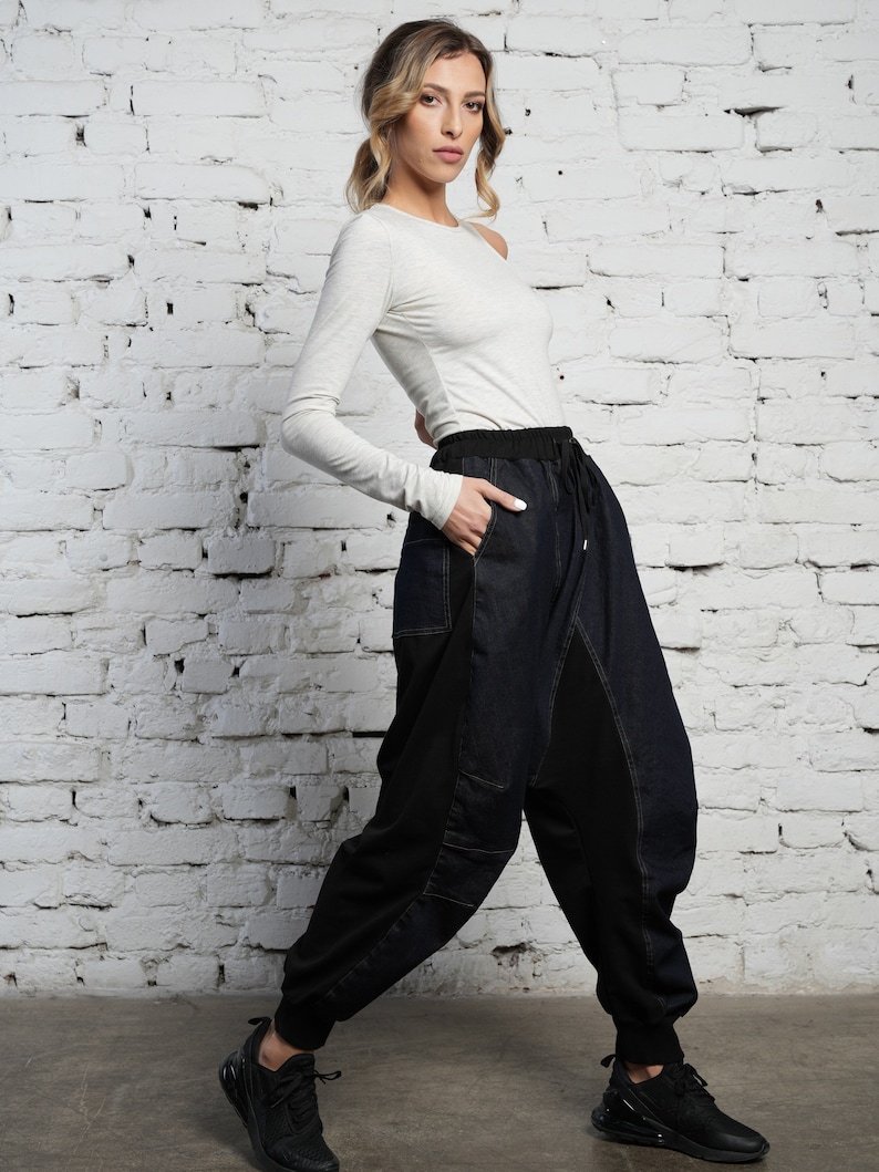 May include: A woman wearing a light gray long-sleeved shirt and dark blue denim pants with black panels. The pants have a drawstring waist and a relaxed fit. She is wearing black sneakers.