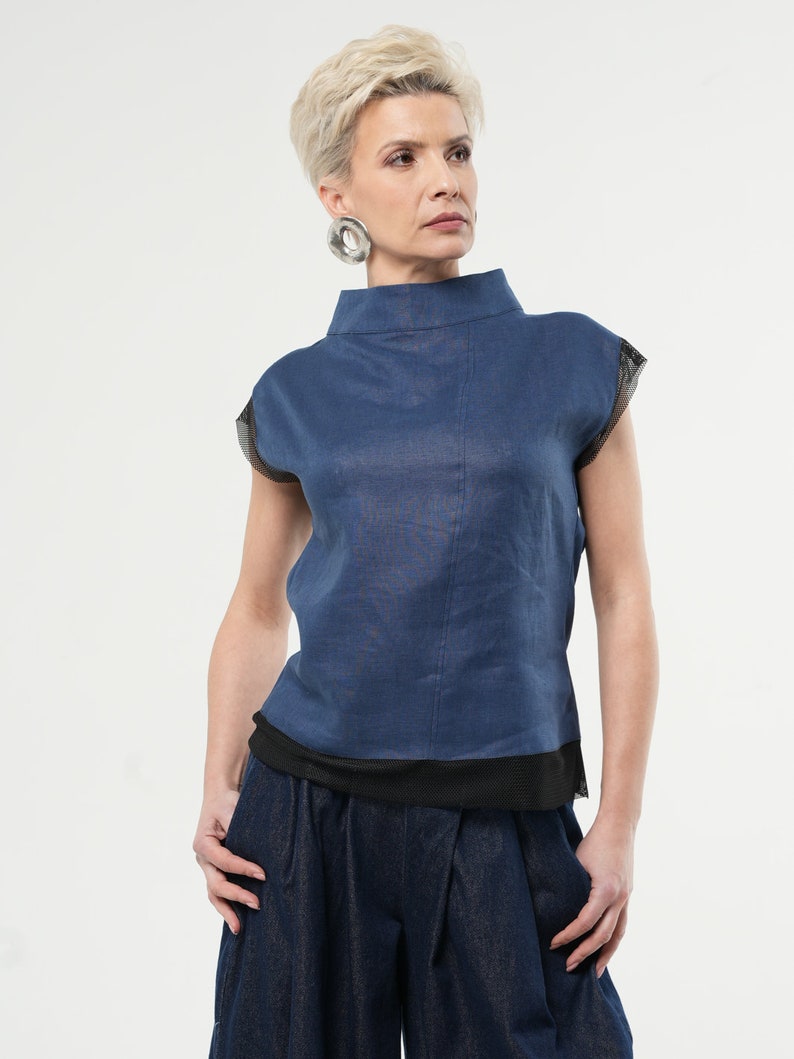 May include: A blue linen top with a mock neck and short sleeves. The sleeves have a black mesh trim. The top is layered over a black mesh tank top. The model is wearing blue denim trousers.