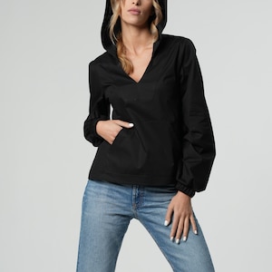 May include: Black hooded pullover shirt with a V-neck and a front pocket. The long-sleeved shirt has elastic cuffs and is paired with light blue denim jeans. The model is wearing the shirt and jeans.