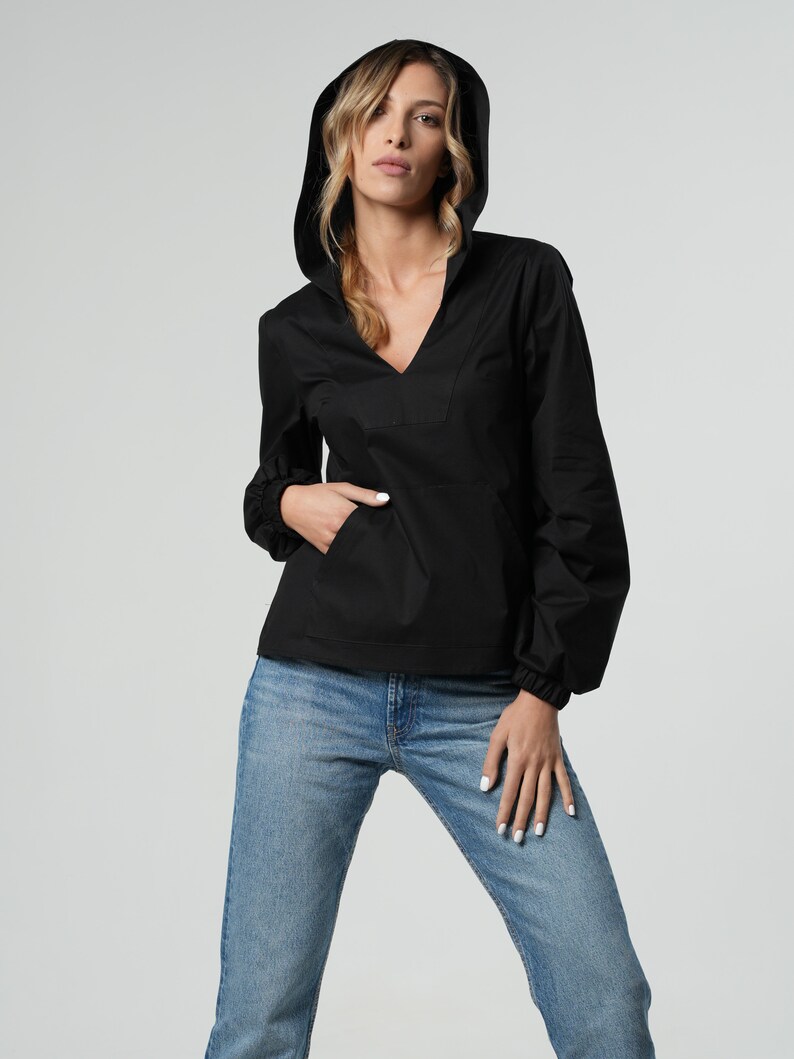 May include: Black hooded pullover shirt with a V-neck and a front pocket. The long-sleeved shirt has elastic cuffs and is paired with light blue denim jeans. The model is wearing the shirt and jeans.