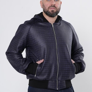 May include: A navy blue bomber jacket with a textured pattern, a hood, and a silver zipper. The jacket has black ribbed cuffs and a black waistband. The person is wearing dark blue jeans.