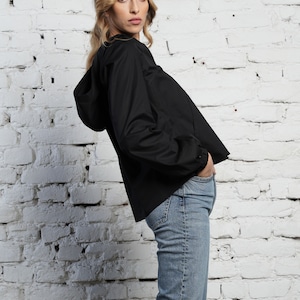 May include: A black hooded jacket with long sleeves and a drawstring hood. The jacket is paired with light blue denim jeans. The model is standing in front of a white brick wall.