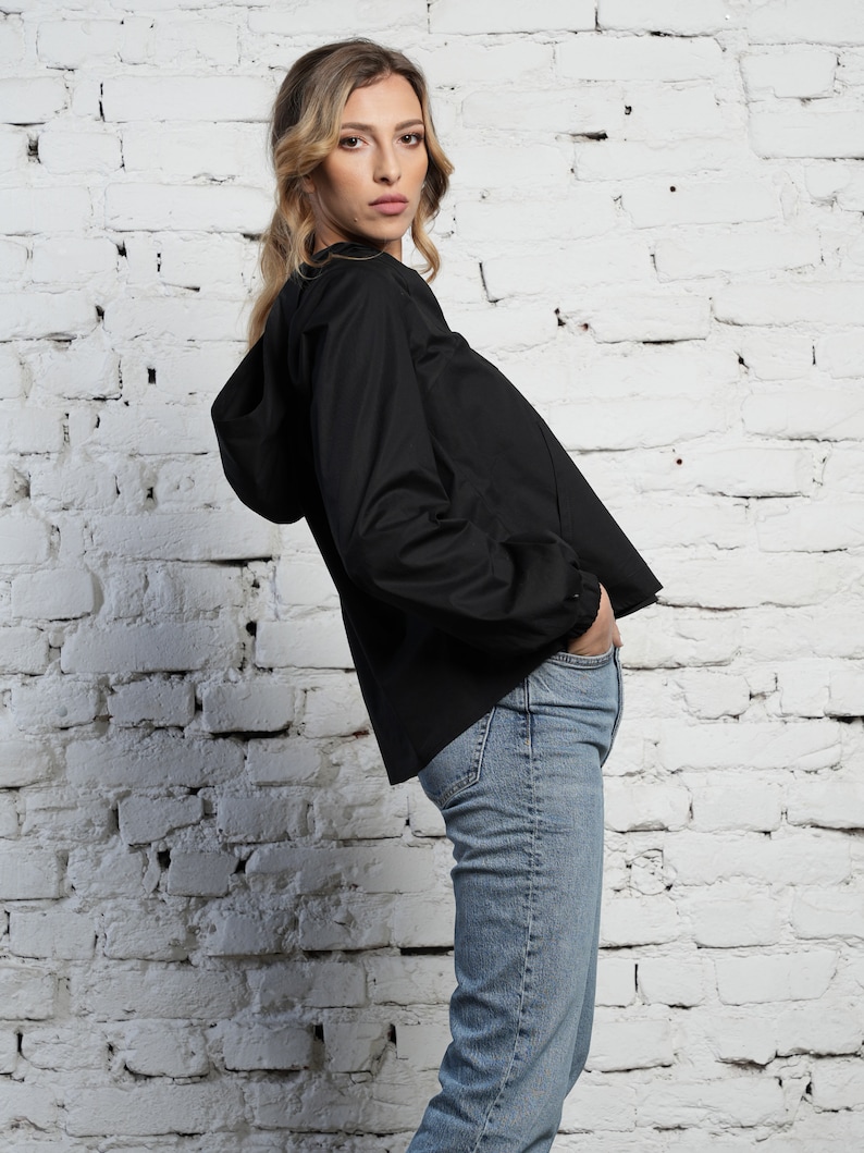May include: A black hooded jacket with long sleeves and a drawstring hood. The jacket is paired with light blue denim jeans. The model is standing in front of a white brick wall.