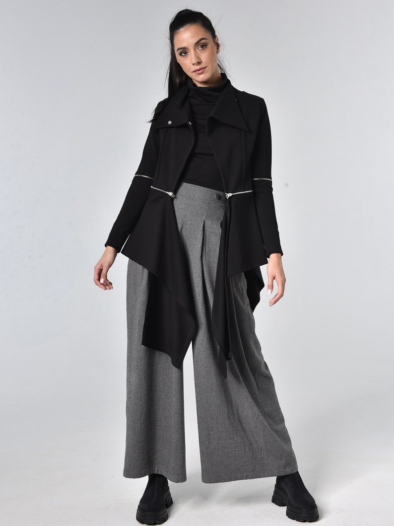 Puede incluir: Una chaqueta negra asim&eacute;trica con detalles de cremallera y cuello alto, combinada con pantalones grises de pierna ancha.