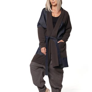 May include: A dark brown open-front cardigan featuring dark blue denim panels on the shoulders and pockets, paired with a matching tie belt. The outfit includes loose-fitting dark brown trousers and light beige loafers.