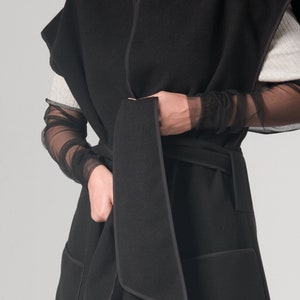 May include: A black sleeveless wrap coat with a self-tie belt and two large pockets. The coat has a contrasting black trim around the edges.