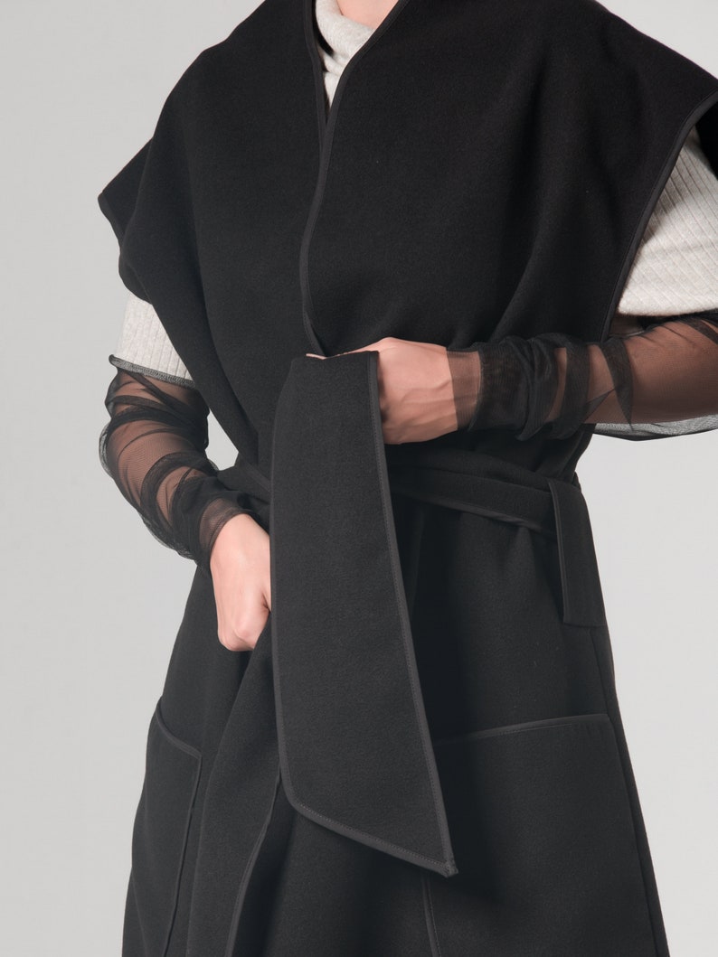 May include: A black sleeveless wrap coat with a self-tie belt and two large pockets. The coat has a contrasting black trim around the edges.