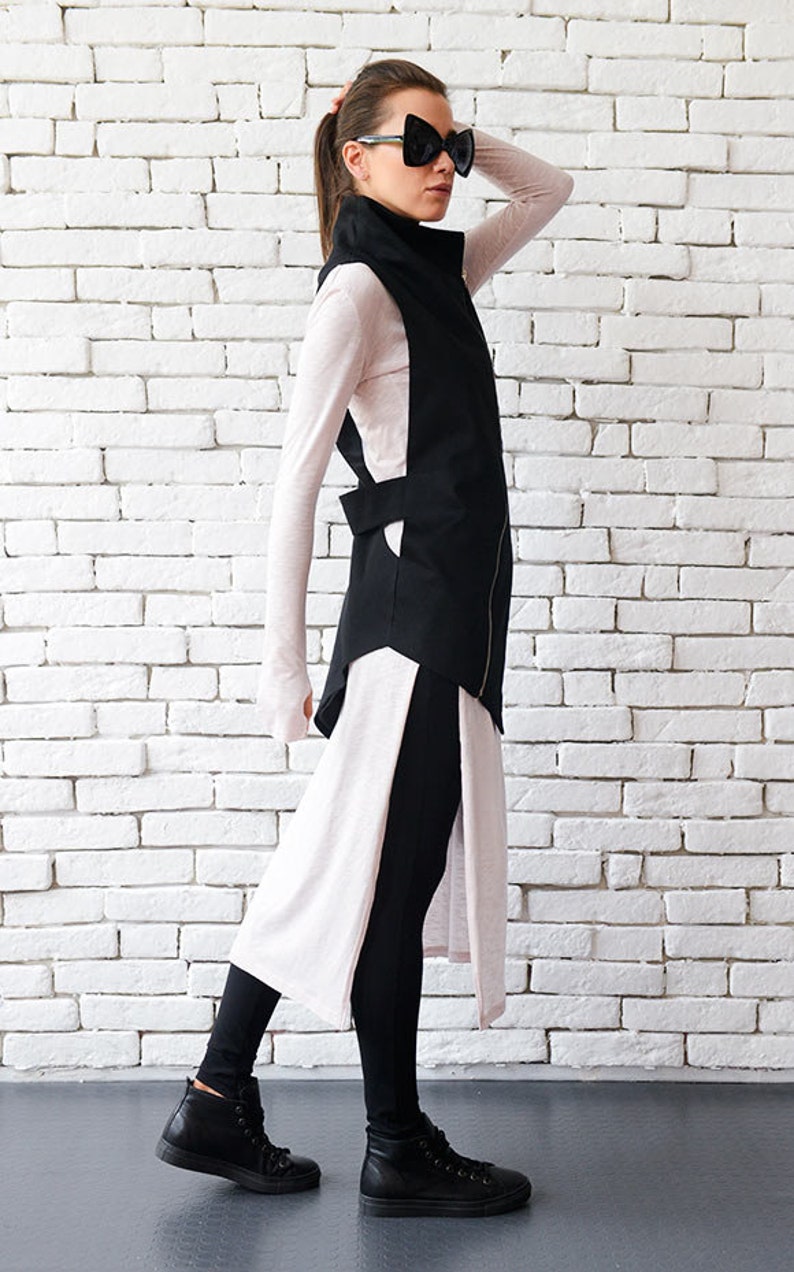 May include: A black long vest with a zipper and a high collar, worn over a pink long-sleeved shirt and black leggings. The person is wearing black high-top sneakers and black sunglasses.