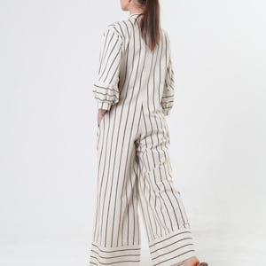 May include: A white and black striped jumpsuit with a wide leg and a cuffed hem. The jumpsuit has a button-up front and a relaxed fit. The model is wearing a brown baseball cap.