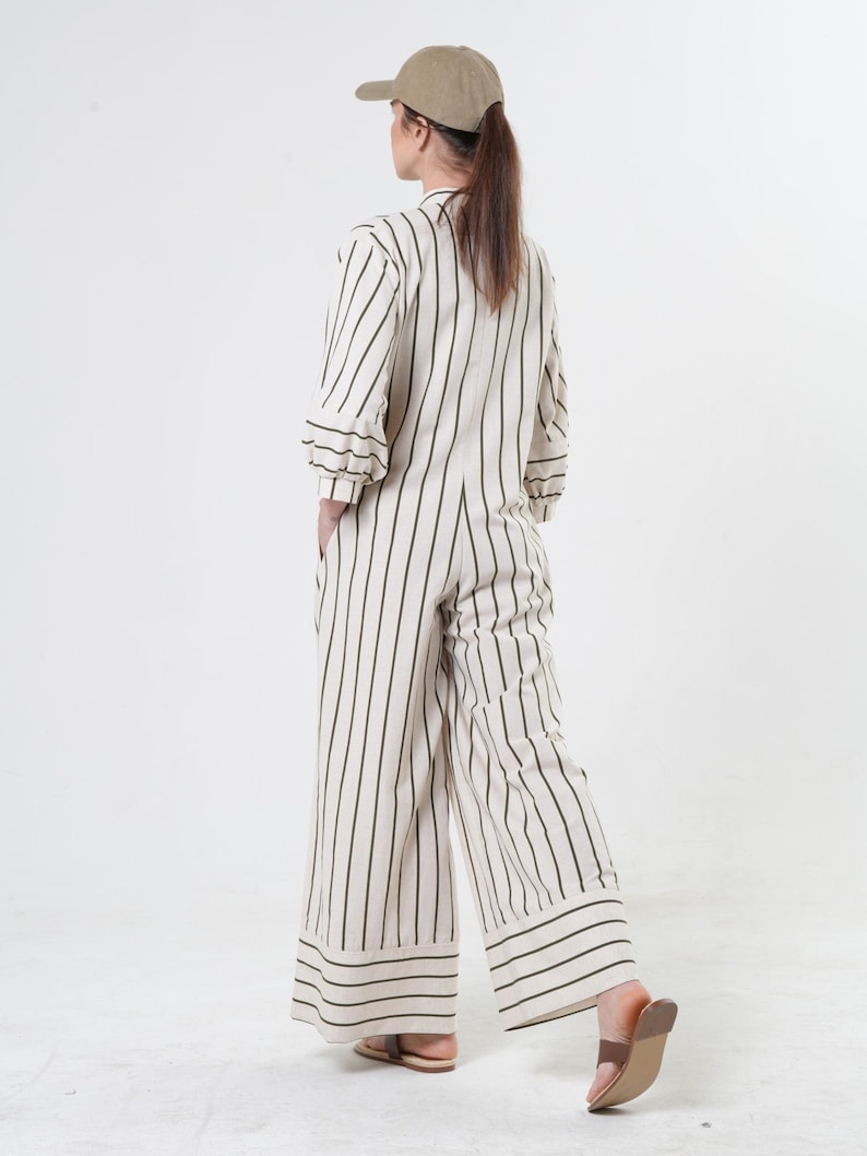 May include: A white and black striped jumpsuit with a wide leg and a cuffed hem. The jumpsuit has a button-up front and a relaxed fit. The model is wearing a brown baseball cap.