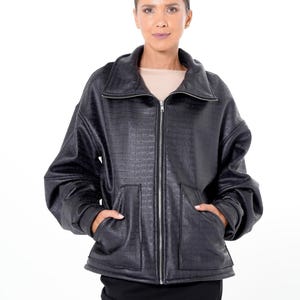 May include: A black faux leather jacket with a textured, crocodile-like pattern. The jacket features a zip-up front, a wide collar, and two front pockets. The sleeves are voluminous, and the jacket is worn over a neutral-colored top and black skirt.