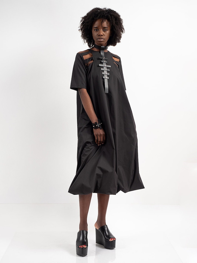 May include: A black midi dress with a cut-out design on the chest and a black leather harness with silver buckles. The dress has a loose fit and a flowing skirt. The model is wearing black platform sandals.