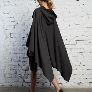 May include: A dark gray hooded cape with a long, asymmetrical hemline. The cape is made of a soft, drapey fabric and has a relaxed fit.