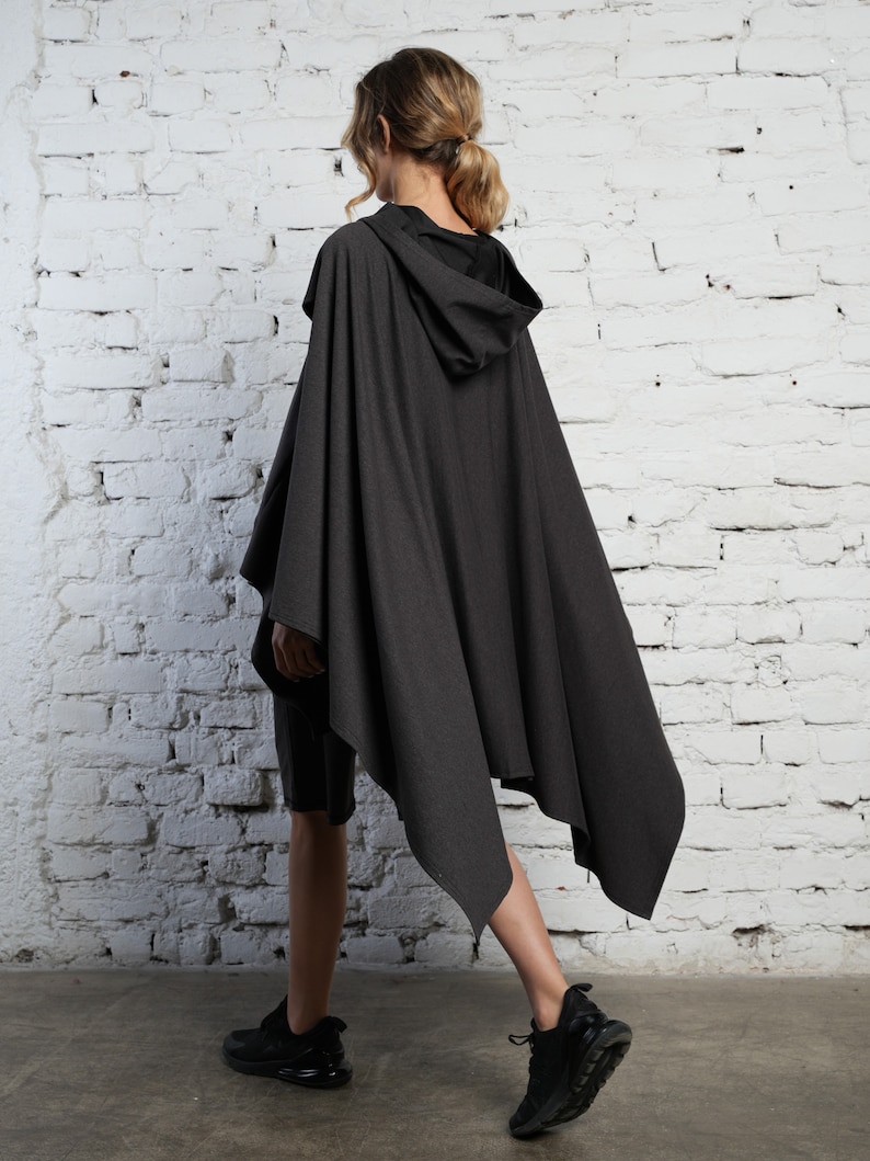 May include: A dark gray hooded cape with a long, asymmetrical hemline. The cape is made of a soft, drapey fabric and has a relaxed fit.