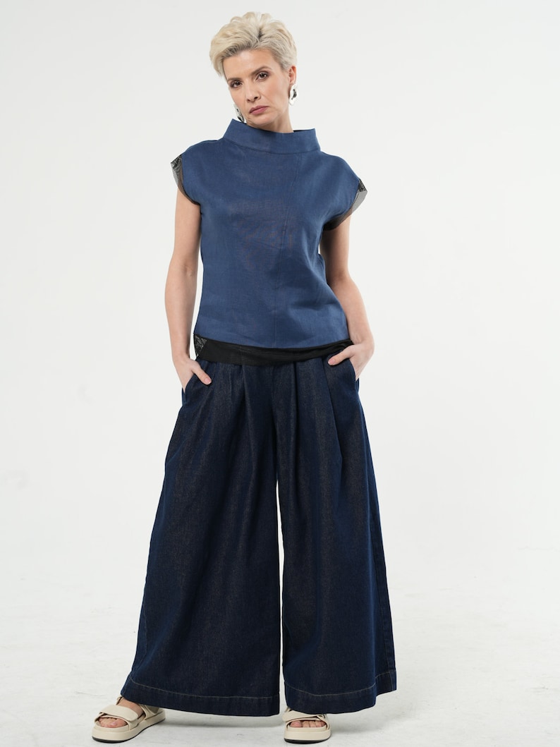 May include: A woman wearing a blue linen top with a high neckline and a pair of dark blue wide-leg denim trousers.