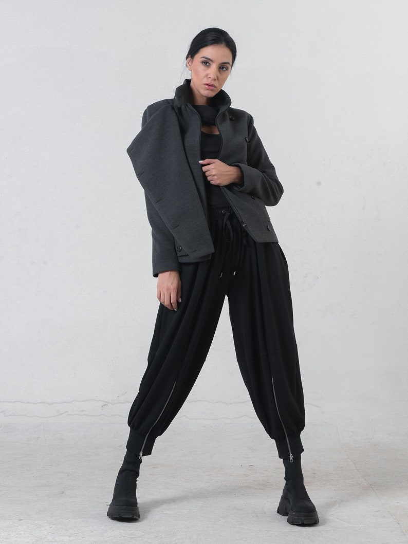 May include: A woman wearing a dark gray jacket with a high collar and black pants. The jacket has button details and a zipper closure. The black pants have a unique, loose fit with zippers at the ankles. The outfit is a modern, stylish ensemble.