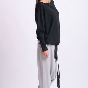 May include: A black long-sleeved top with a boat neck, paired with light gray harem pants and black sneakers. The outfit includes a black belt with a long, dangling strap. The model is wearing black leggings under the pants.