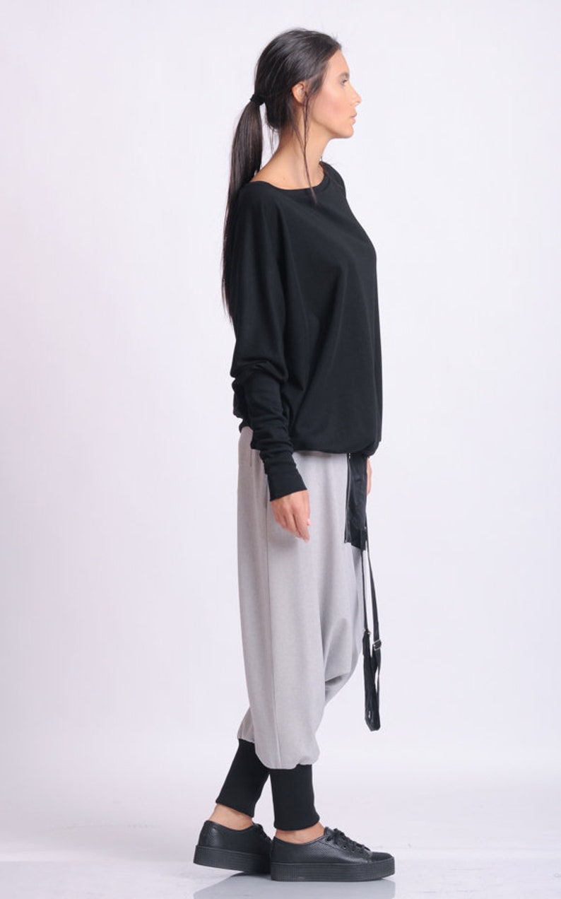 May include: A black long-sleeved top with a boat neck, paired with light gray harem pants and black sneakers. The outfit includes a black belt with a long, dangling strap. The model is wearing black leggings under the pants.