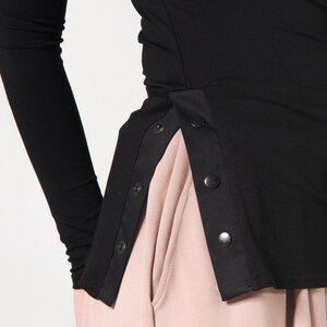 May include: Black long-sleeved shirt with a side panel of black fabric and snap buttons. The shirt is worn with a light pink skirt.