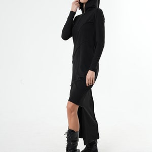 May include: A black long-sleeved hooded dress with a high side slit. The dress has a fitted silhouette and a relaxed fit around the shoulders. The model is wearing black boots.