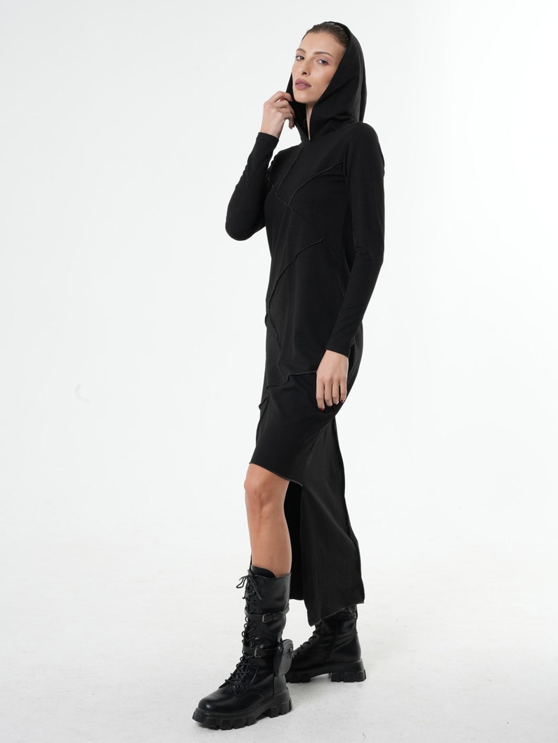 May include: A black long-sleeved hooded dress with a high side slit. The dress has a fitted silhouette and a relaxed fit around the shoulders. The model is wearing black boots.